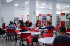 Interior da biblioteca da universidade. Mesas redondas com cadeiras vermelhas acomodam estudantes que leem e fazem pesquisas. Ao fundo se observa as prateleiras de livros.