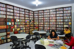 Sala com quatro mesas redondas onde se vê três acadêmicas. Duas delas dividem uma das mesas com vários livros. Ao fundo uma acadêmica usa uma mesa e sobre ela um notebook. Nas duas paredes da sala há prateleiras do teto ao chão com DVDs de filmes e alguns livros dispostos de forma organizada.