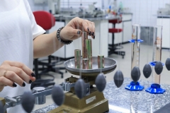 Detalhe de duas mãos de uma mulher, com as unhas pintadas, fazendo um experimento dentro de um laboratório da universidade, com uma balança.