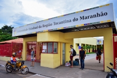 Fachada externa da universidade. Dois portões grandes, um deles está aberto. Entre os portões uma guarita. Sobre os portões e a guarita, no alto o nome: Universidade Estadual da Região Tocantina do Maranhão. Em frente ao portão estão três pessoas. No meio-fio um motoqueiro do serviço de moto-táxi espera um passageiro.