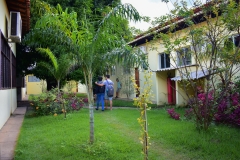 Área interna da universidade entre dois blocos, com ajardinamento onde se vê flores, gramado e alguns arbustos. Um casal caminha abraçado pelo gramado, tendo mochilas nas costas.