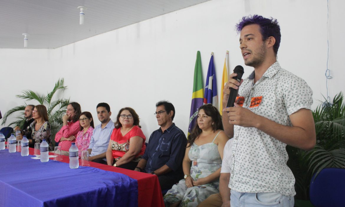 Um estudante, que tem adesivos do grupo Juventude Anticapitalista colados em sua camisa, fala ao microfone ao lado da mesa da gestão superior da UEMASUL.