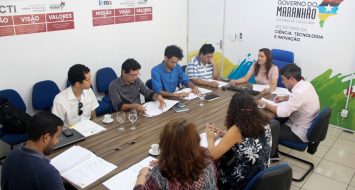 Um grupo de pessoas aparece sentado ao redor de uma mesa em reunião.