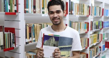 A foto tem como fundo prateleiras de livros da biblioteca da UEMASUL. O historiador Wekslley Machado mostra o livro de sua autoria: Templários: Entre a pureza e a sodomia no século XIV.