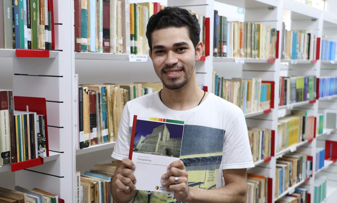 A foto tem como fundo prateleiras de livros da biblioteca da UEMASUL. O historiador Wekslley Machado mostra o livro de sua autoria: Templários: Entre a pureza e a sodomia no século XIV.