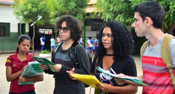 Na foto, um grupo de jovens acadêmicos está reunido no pátio de uma escola, conversando. Ao fundo se vê algumas árvores. Dois estudantes estão de costas, e os outros quatro de frente.