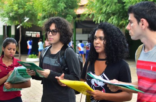 Na foto, um grupo de jovens acadêmicos está reunido no pátio de uma escola, conversando. Ao fundo se vê algumas árvores. Dois estudantes estão de costas, e os outros quatro de frente.