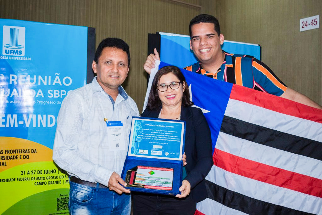 Orientador Zilmar Timóteo e orientando comemoram premiação. Reitora Elizabeth Nunes Freire e orientador seguram premiações. Orientando, Brunno Gustavo segura bandeira do Maranhão representando os discentes do curso.