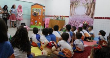 Crianças de escolas municipais reunidas em uma sala e sentadas em um tapete vermelho. A frente temos os acadêmicos vestidos com fantasias de personagens infantis.