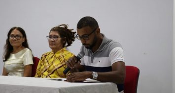 Sentados à mesa do auditório da UEMASUL, à direita duas professoras, à esquerda o acadêmico, com microfone na mão, lendo seu discurso.