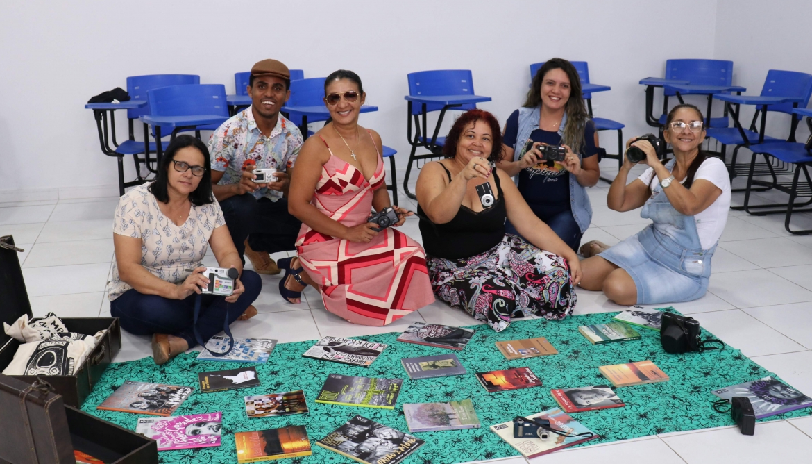 Seis professores sentados no chão, em uma sala de aula, seguram objetos e livros usados na formação.