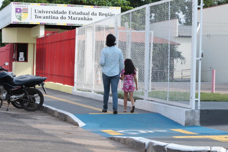 Uma mulher adulta caminha pela calçada tátil da UEMASUL de mãos dadas com uma criança.