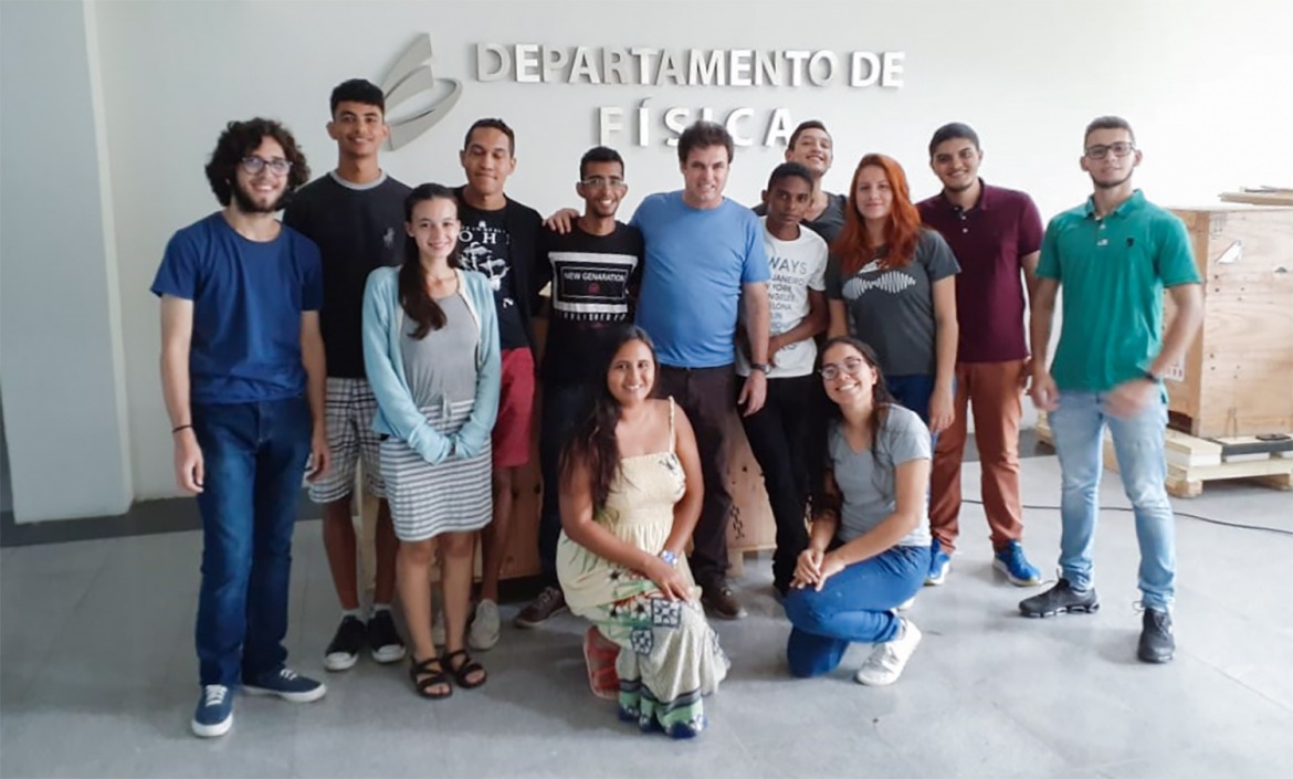 13 estudantes posam para foto. Duas estão agachadas. Atrás deles na parede, o nome: Departamento de Física.