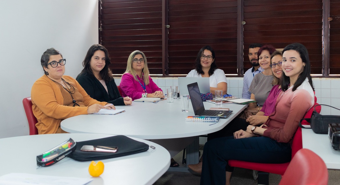 Oito pessoas sentadas à mesa de reunião, posam para foto e sorriem.